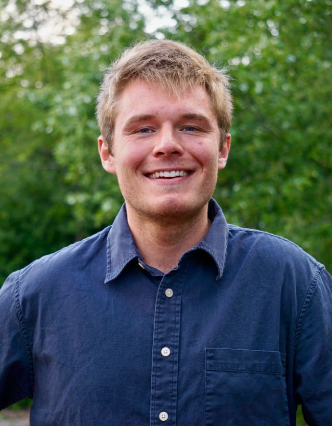 Man with blonde hair wearing a dark blue shirt smiling standing in front of green trees