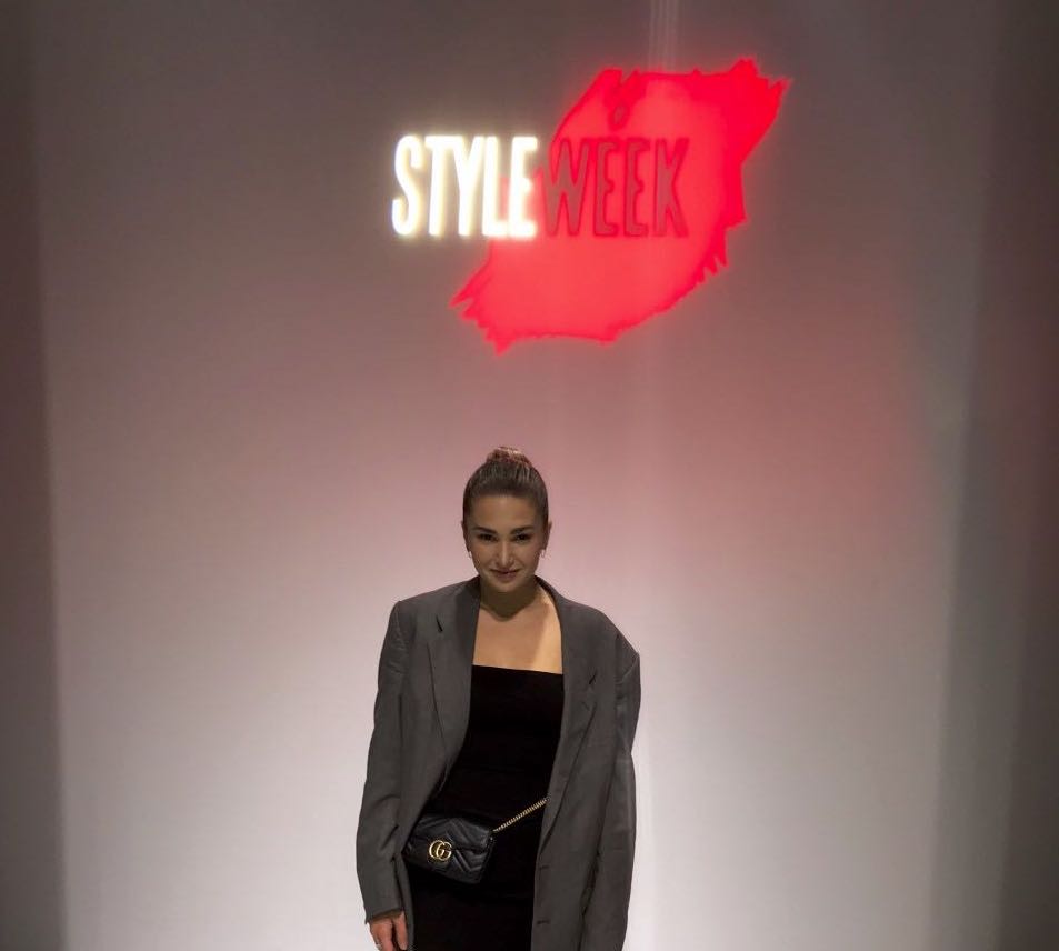 Photo of person wearing a black dress with a grey blazer and a black purse standing in front of a wall that says StyleWeek 