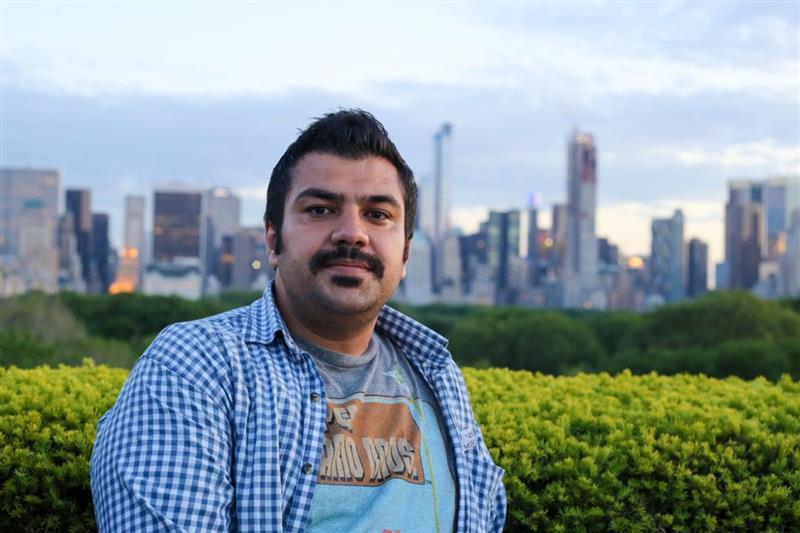 Man with black hair sitting in front of a city skyline. He is wearing a checkered button up with a graphic tshirt underneath