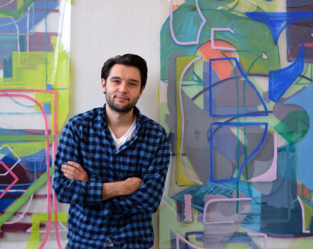 Man with black hair standing in front of a colorful art exhibit. He is wearing a blue and black checkered shirt.