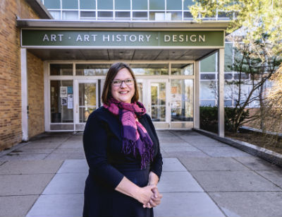 Woman wearing a black sweater and pink and purple scarf. She is standing in front of MSU's Art, Art History, and Design