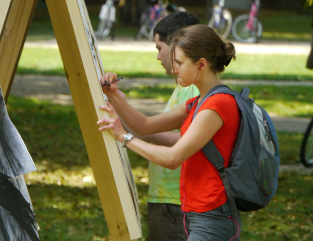 A woman in the foreground is wearing a red t-shirt and backpack. She is drawing on an easel with a man in the background, who is wearing a green shirt