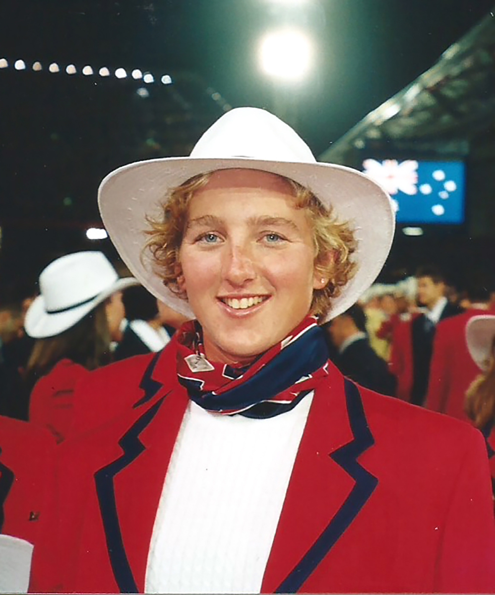 A young girl with blonde hair and blue eyes smiling. She is wearing a white coyboy styled hat and a bandana around her beck and a red blazer.