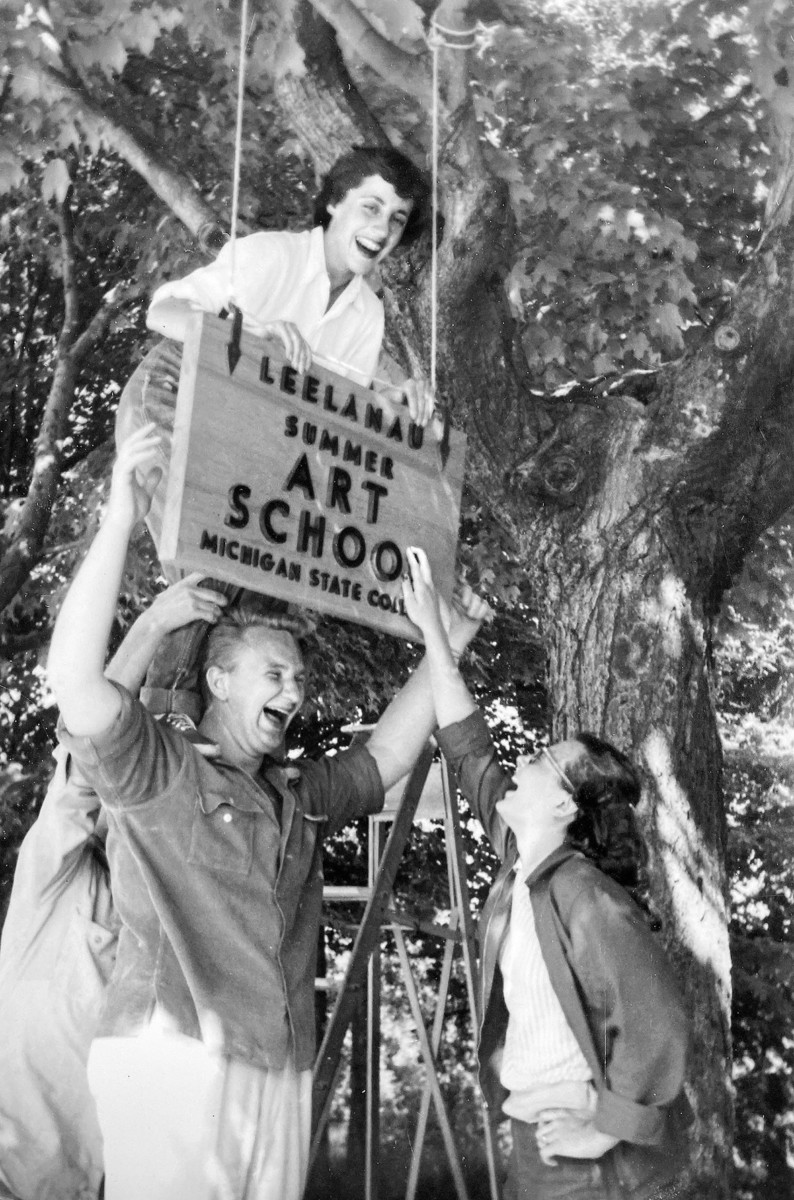 Black and white photograph of a woman standing on a man's shoulders hanging up a wooden sign that says "Leland Summer Art School, Michigan State College"