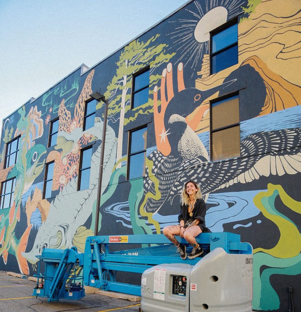 Smiling young woman sits on a blue power lift machine in front of a mural featuring animals and nature.