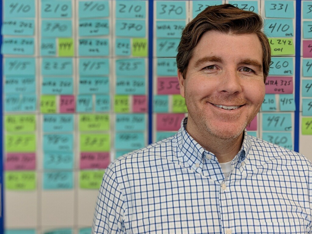 Man with red hair wearing a blue and white shirt. Behind him is a whiteboard filled with blue, green, and pink sticky notes with numbers written on them.