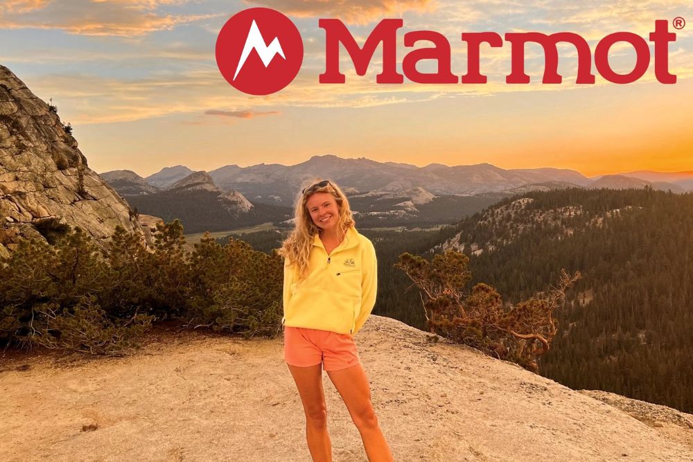 A young woman with blonde hair wearing a yellow sweater and orange shorts stands in front of a mountain landscape at sunset.