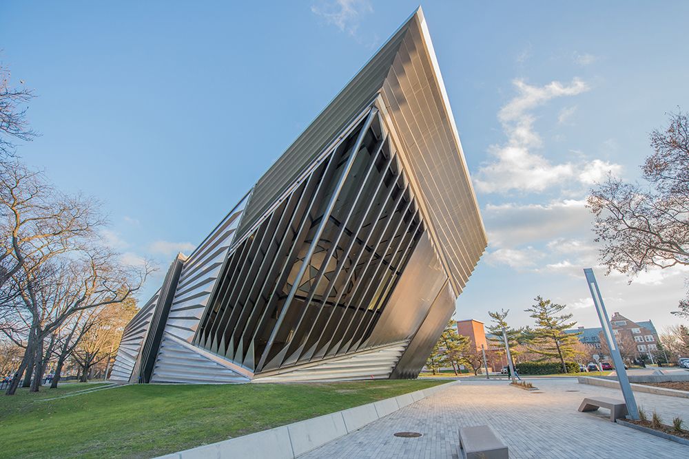 A building made of diagonal steel panels in alternating directions sits on a university campus surrounded by blue skies, gray sidewalks, and green grass.