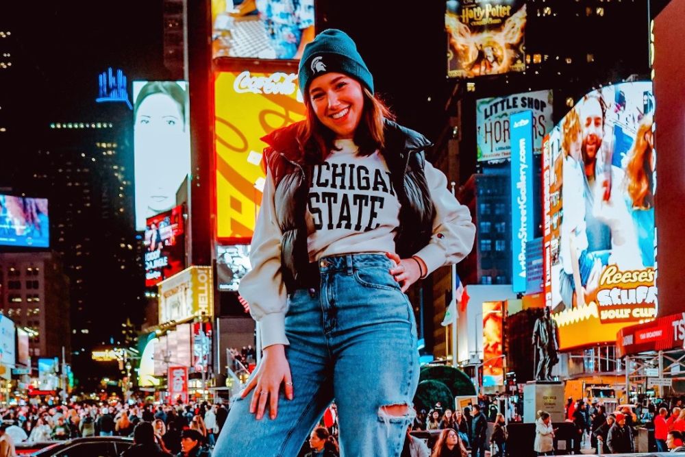 A young smiling woman stands in front of the tall buildings and bright lights of Times Square.