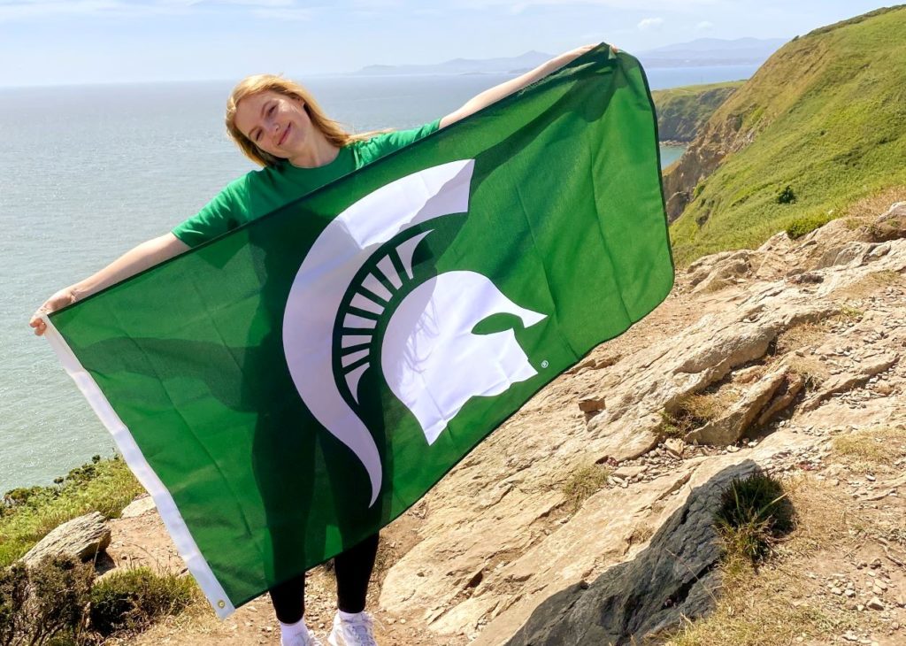 Person standing on cliff with water in the background. The person is holding a green flag with a Spartan logo.