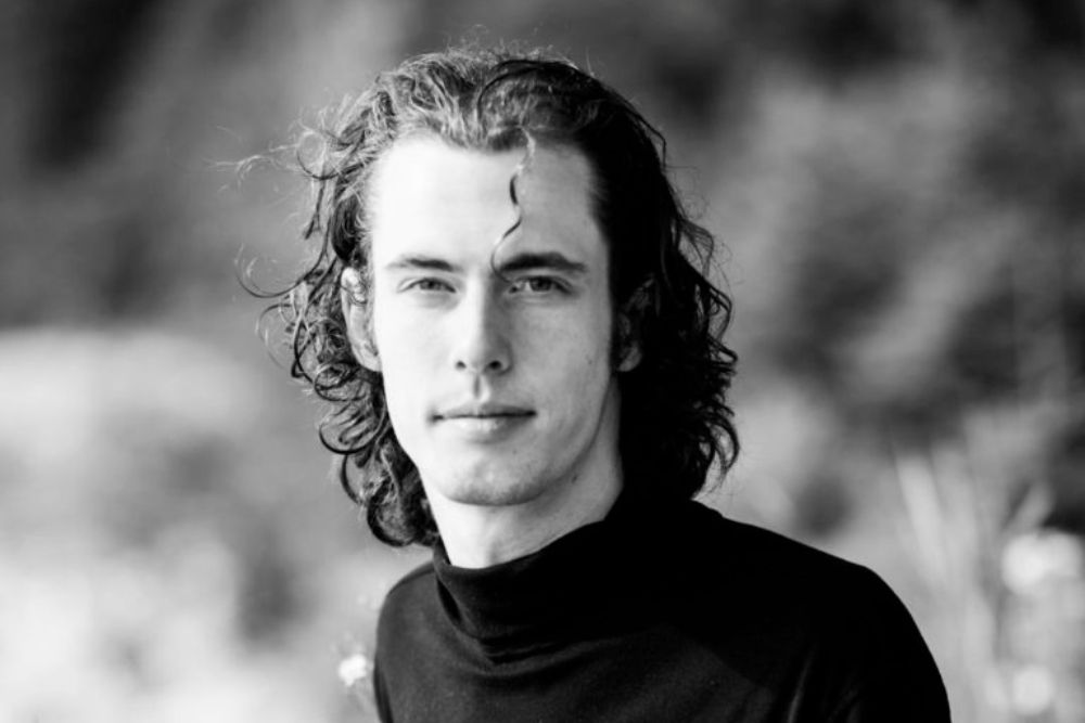 A black-and-white portrait of a young man with curly hair wearing a black turtleneck.