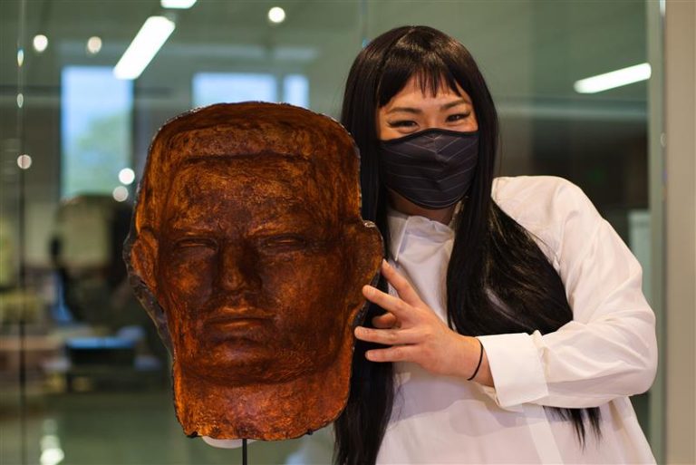 Photo of a person standing next to a metal casting of a head. They have long black hair and are wearing a face mask and white dress shirt.