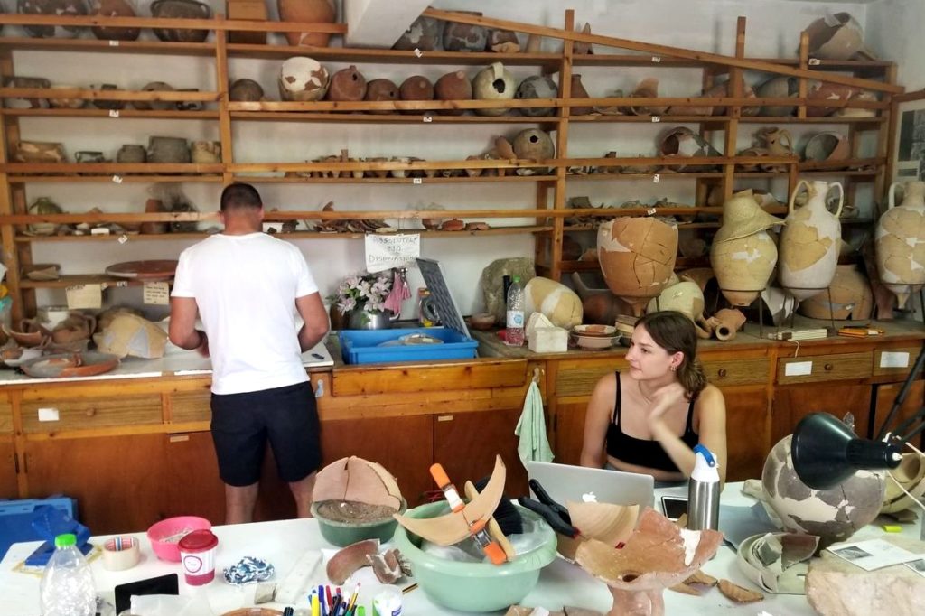 Photo in a room with several ancient artifacts all around with a man who is standing and has his back turned and a woman who is sitting and working at a computer. 