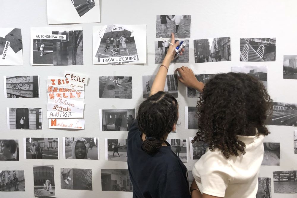 Photos and collages with words are posted up on a wall. Two adolescents look at the exhibit and point at some of the displayed images.