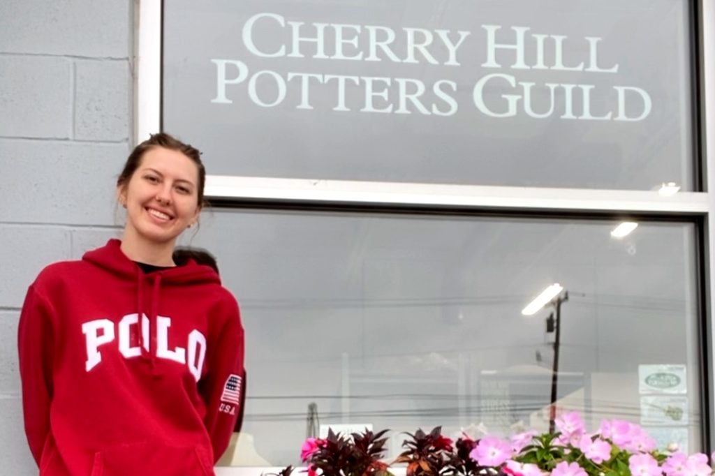 Woman with hair pulled back into a ponytail wearing a red hooded sweatshirt that says "Polo" on it standing in front of a window with the words "Cherry Hill Potters Guild" 