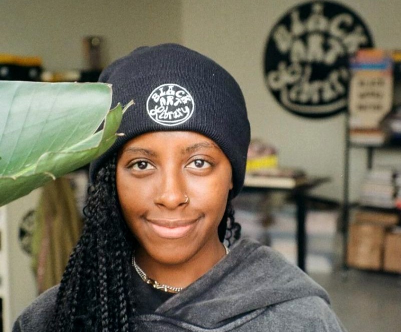 Black woman wearing a black knit hat that says "Black Art Library" on the front. 