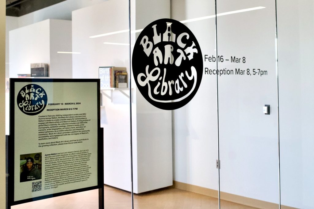 A glass wall with the "Black Art Library" logo on it and the dates Feb. 16-Mar 8, 5-7 p.m. Standing by this wall is a sign with more information about the exhibit. 