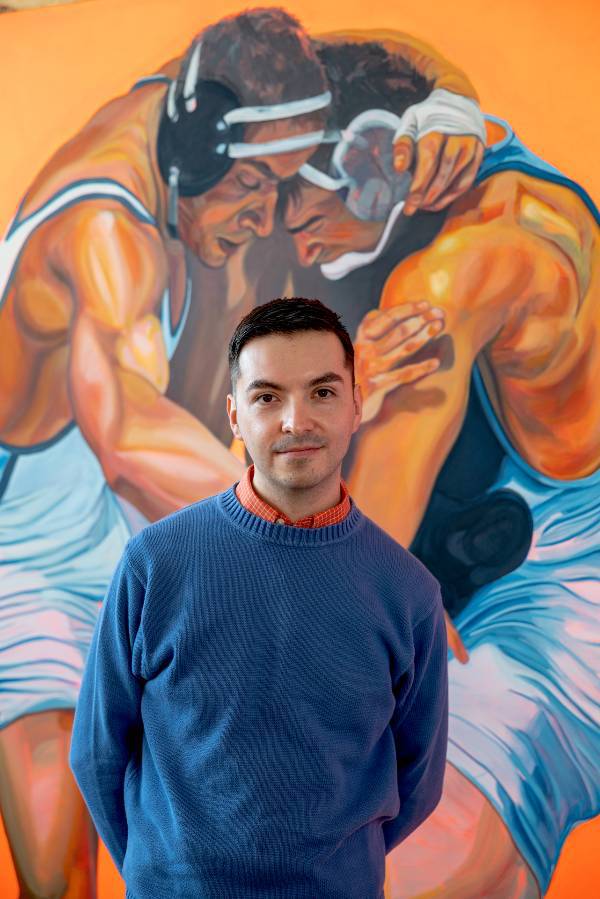 Gustavo Uriel Ayala standing in front of one of his paintings depicting two male wrestlers. 