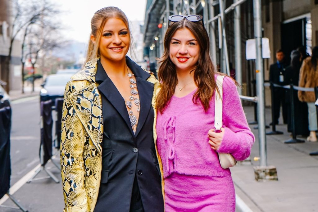 Two women side by side. On the left is a person in a gold snakeskin jacket and a black suit jacket, on the right is a person in a bright pink sweater and skirt carrying a white purse.