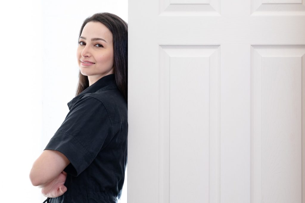 Woman with long dark hair earing a black shirt leaning up agains a white door with a white background.