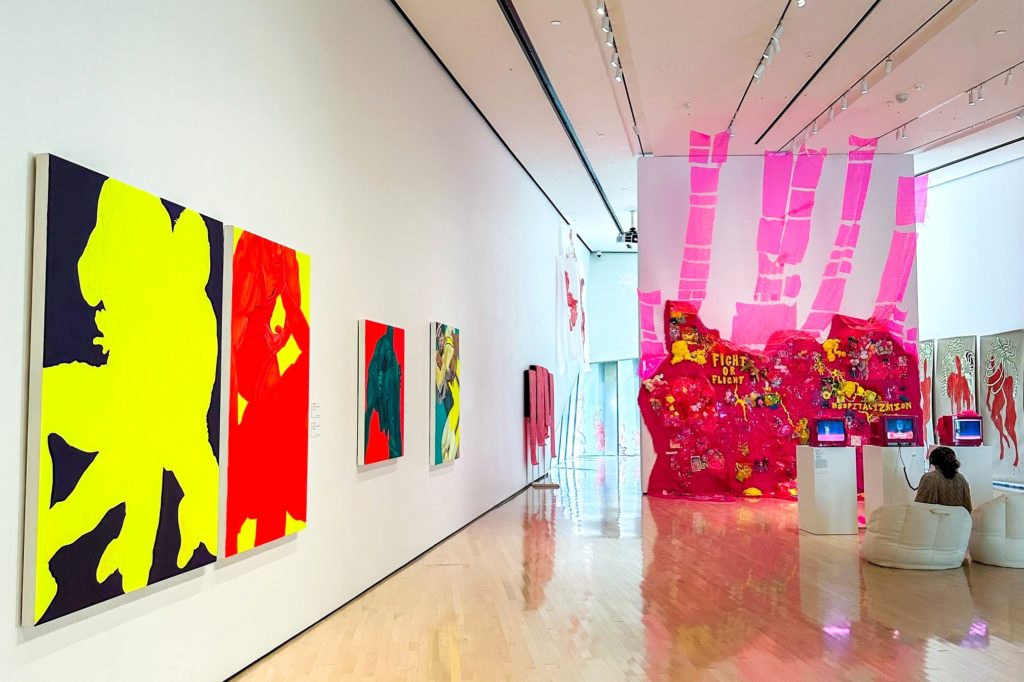 Photo inside an art gallery with four brightly colored paintings hanging on the wall on the left side of the photo and a bright pink piece of art on the right side of the photo with a person, whose back is to the camera, sitting in a chair looking at the bright pink piece. 
