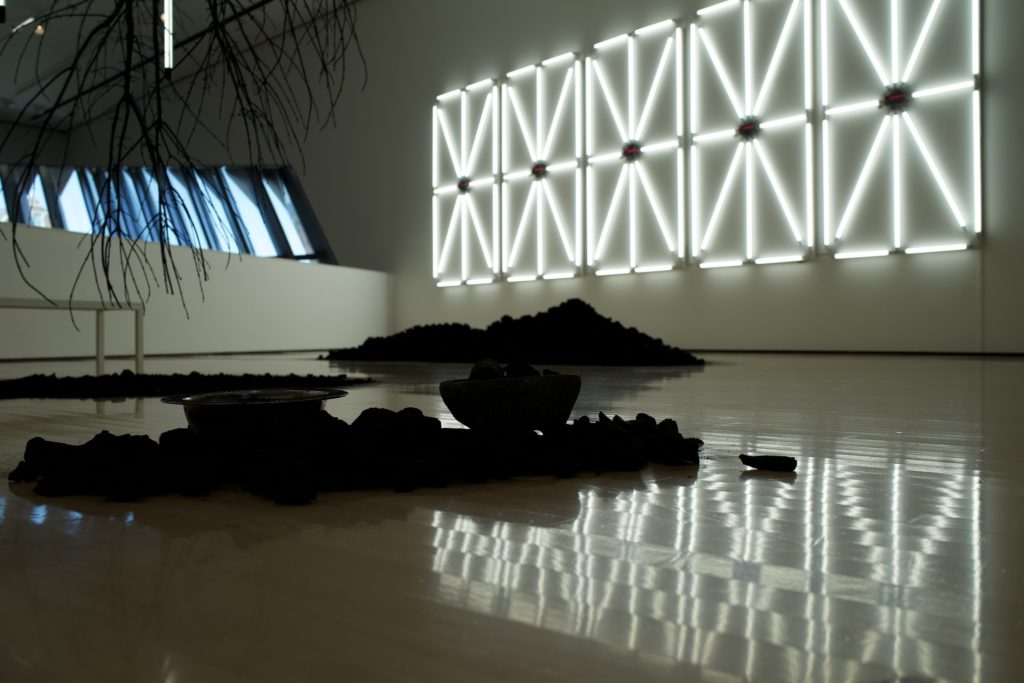 A picture of an art installation. It is a tree in front of a wall of white neon lights. There are some stones and a bowl on the ground.