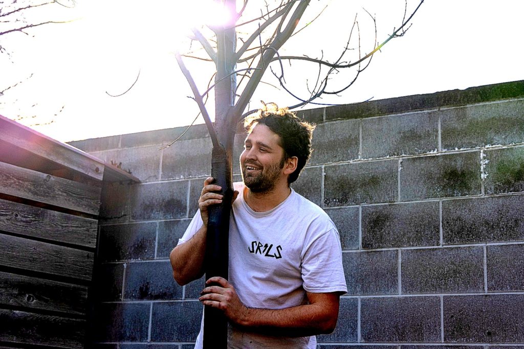Photo of a man with dark brown hair, a beard, and a mustache, wearing a white T-shirt and holding onto the truck of a small tree trunk.
