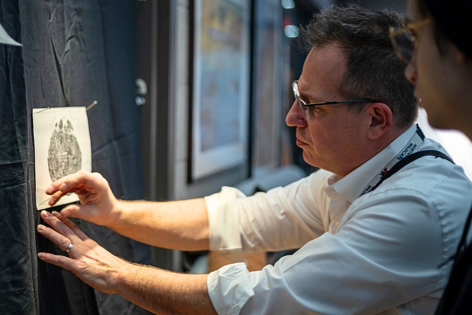 A picture of a man wearing a white button-down shirt in front of a sketch on white paper. A person with glasses watches him work