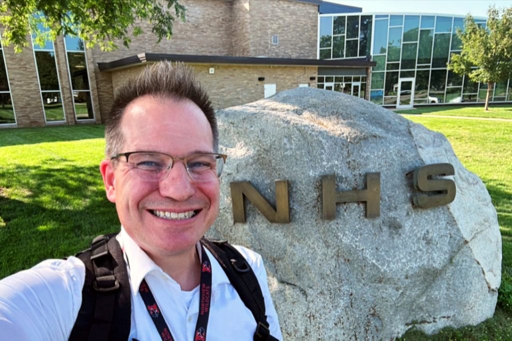 A picture of a man with short hair and glasses, wearing a white button down and backpack, in front of a rock with NHS on it and a large building. 