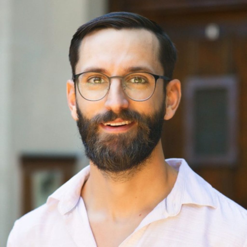 Portrait of a smiling man with dark hair and beard wearing eyeglasses and a white button-down.