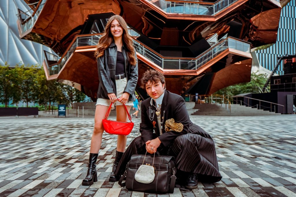 A photo of two people standing in front of an architectural display. The woman on the left has long brown wears a black leather jacket and carries a red purse; the man on the right has short, choppy hair and wears a suit coat covered in pins and a dress. 