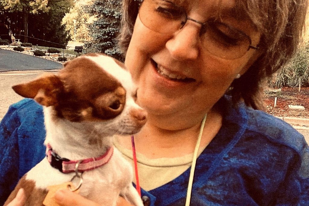 Woman wearing glasses and a dark blue shirt holding and looking at a small brown and white dog.